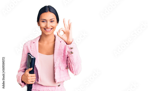Young beautiful latin girl wearing business clothes holding binder doing ok sign with fingers, smiling friendly gesturing excellent symbol