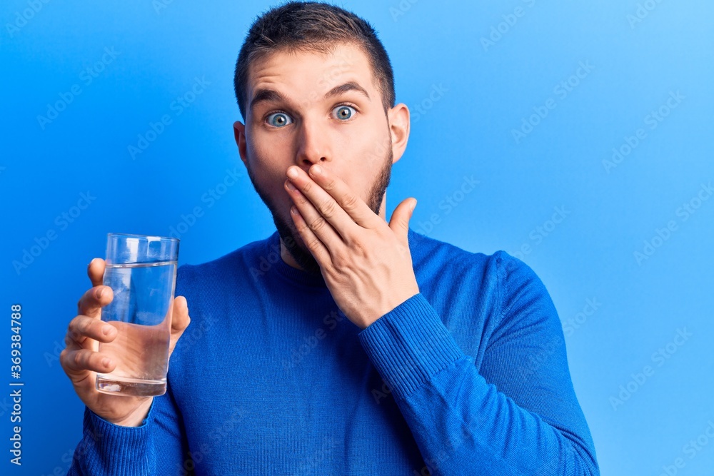 Young handsome man drinking glass of water covering mouth with hand, shocked and afraid for mistake. surprised expression Young handsome man drinking glass of water covering mouth with hand, shocked and afraid for mistake. surprised expression