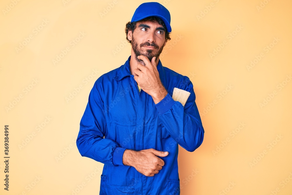 Handsome young man with curly hair and bear wearing builder jumpsuit uniform with hand on chin thinking about question, pensive expression. smiling with thoughtful face. doubt concept.