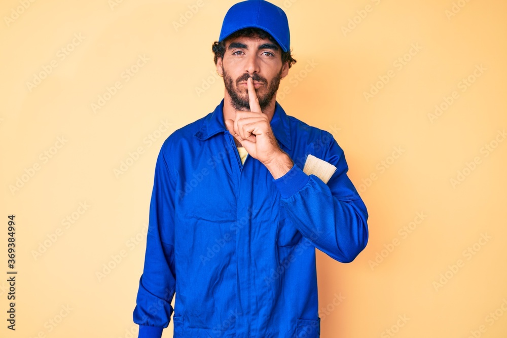 Handsome young man with curly hair and bear wearing builder jumpsuit uniform asking to be quiet with finger on lips. silence and secret concept.