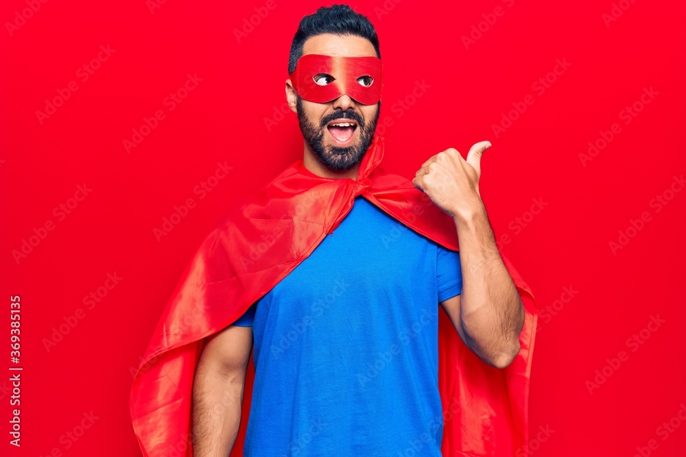 Young hispanic man wearing super hero costume pointing thumb up to the side smiling happy with open mouth