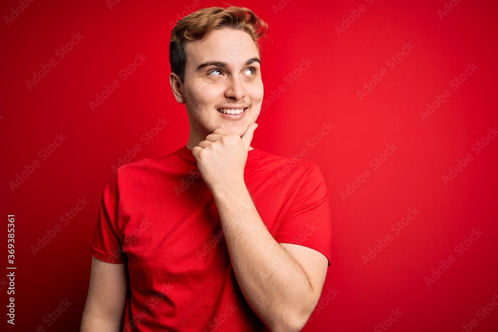 Young handsome redhead man wearing casual t-shirt over isolated red background with hand on chin thinking about question, pensive expression. Smiling with thoughtful face. Doubt concept. Young handsome redhead man wearing casual t-shirt over isolated red background with hand on chin thinking about question, pensive expression. Smiling with thoughtful face. Doubt concept.