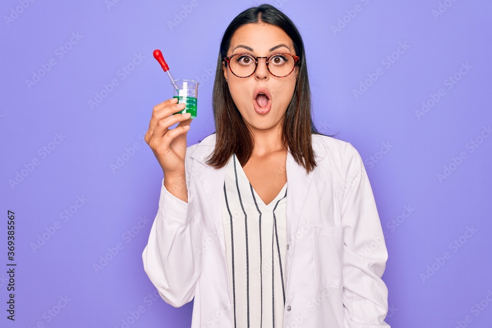 Young beautiful brunette scientist woman wearing coat and glasses holding test tube scared and amazed with open mouth for surprise, disbelief face Young beautiful brunette scientist woman wearing coat and glasses holding test tube scared and amazed with open mouth for surprise, disbelief face