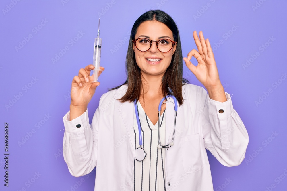 Young beautiful brunette doctor woman wearing stethoscope holding syringe vaccination doing ok sign with fingers, smiling friendly gesturing excellent symbol Young beautiful brunette doctor woman wearing stethoscope holding syringe vaccination doing ok sign with fingers, smiling friendly gesturing excellent symbol