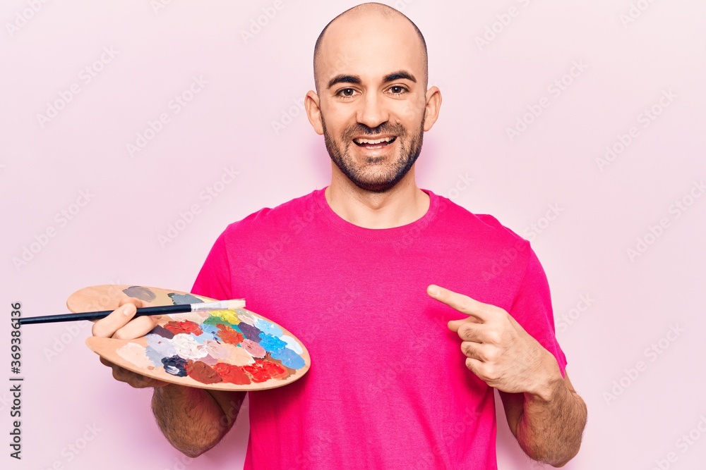 Young handsome bald man painting using paintbrush and palette smiling happy pointing with hand and finger Young handsome bald man painting using paintbrush and palette smiling happy pointing with hand and finger