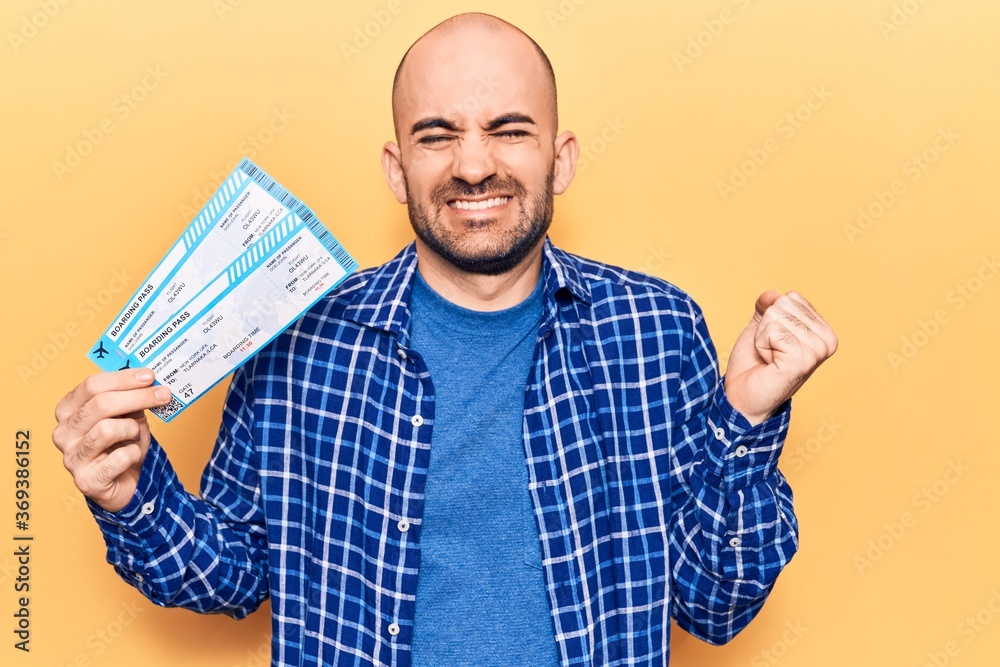 Young handsome bald man holding airline boarding pass screaming proud, celebrating victory and success very excited with raised arm Young handsome bald man holding airline boarding pass screaming proud, celebrating victory and success very excited with raised arm