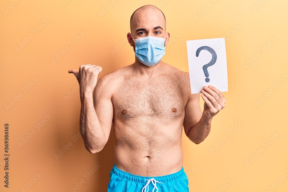 Fototapeta premium Young handsome bald man wearing swimwear and medical mask holding question mark pointing thumb up to the side smiling happy with open mouth