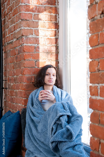 A girl with tea wrapped in a blanket sits on the windowsill near the window in autumn, the concept of comfort and warmth