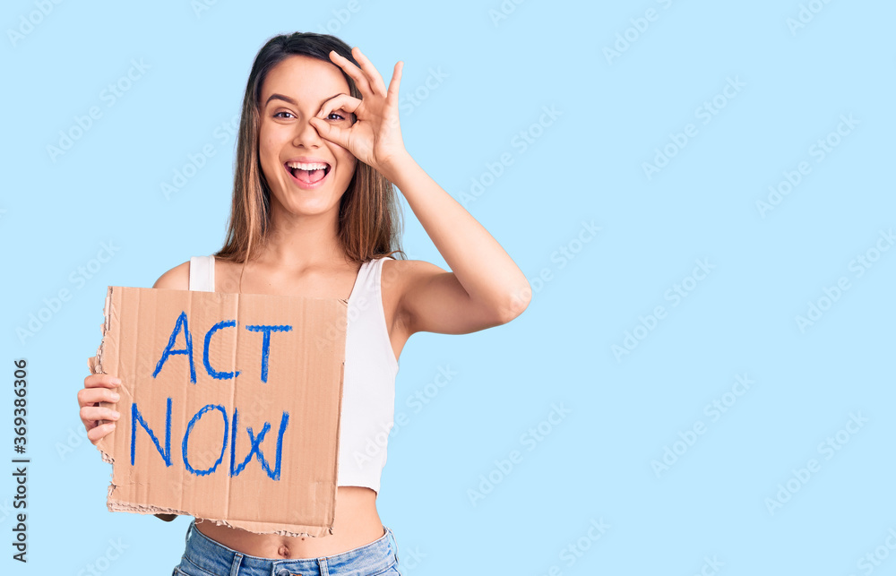 Young beautiful girl holding act now banner smiling happy doing ok sign with hand on eye looking through fingers Young beautiful girl holding act now banner smiling happy doing ok sign with hand on eye looking through fingers