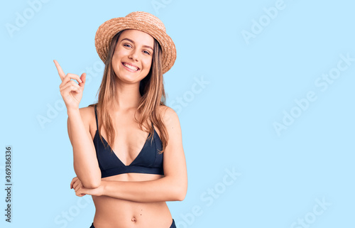 Young beautiful girl wearing bikini and hat with a big smile on face, pointing with hand and finger to the side looking at the camera.