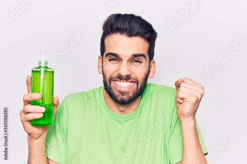 Young handsome man with beard wearing bottle of aloe vera cream screaming proud, celebrating victory and success very excited with raised arm