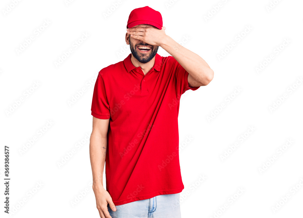 Young handsome man with beard wearing delivery uniform smiling and laughing with hand on face covering eyes for surprise. blind concept. Young handsome man with beard wearing delivery uniform smiling and laughing with hand on face covering eyes for surprise. blind concept.