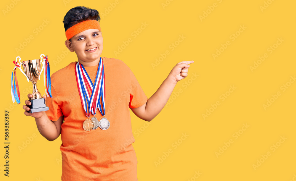 Little boy kid holding champion trophy and wearing medals smiling happy pointing with hand and finger to the side