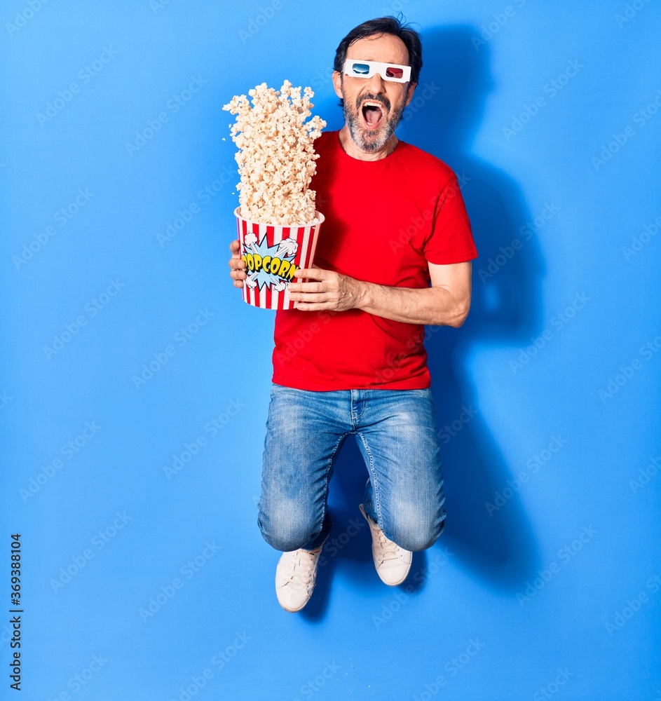 Middle age handsome hispanic man suprised with open mouth watching movie using 3d glasses. Holding bucket of popcorn jumping over isolated blue background Middle age handsome hispanic man suprised with open mouth watching movie using 3d glasses. Holding bucket of popcorn jumping over isolated blue background