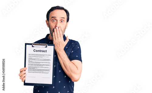 Middle age handsome man holding clipboard with contract document covering mouth with hand, shocked and afraid for mistake. surprised expression photo