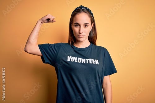 Young beautiful woman wearing volunteer t-shirt doing volunteering over yellow background Strong person showing arm muscle, confident and proud of power