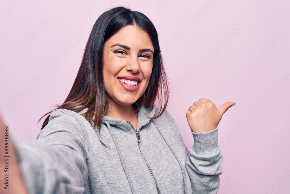 Young beautiful brunette sporty woman make selfie by the camera over pink background pointing thumb up to the side smiling happy with open mouth Young beautiful brunette sporty woman make selfie by the camera over pink background pointing thumb up to the side smiling happy with open mouth