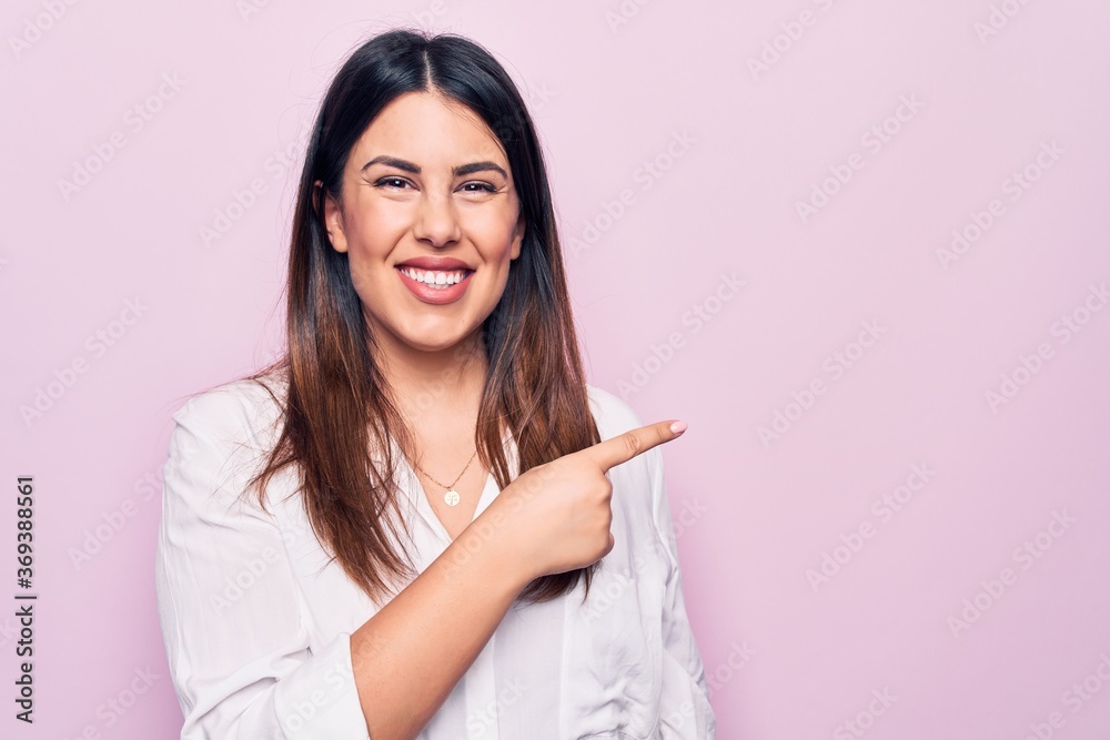 Young beautiful brunette woman wearing elegant shirt standing over isolated pink background smiling cheerful pointing with hand and finger up to the side