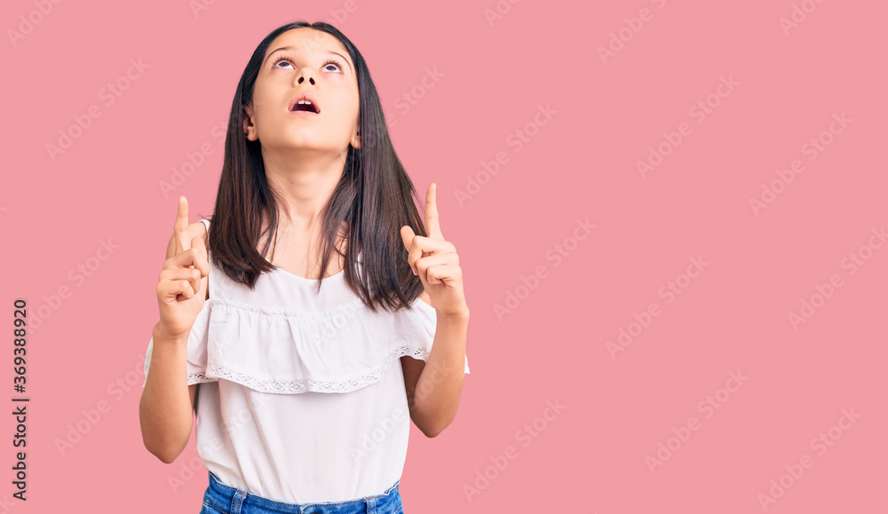 Beautiful child girl wearing casual clothes amazed and surprised looking up and pointing with fingers and raised arms. Beautiful child girl wearing casual clothes amazed and surprised looking up and pointing with fingers and raised arms.
