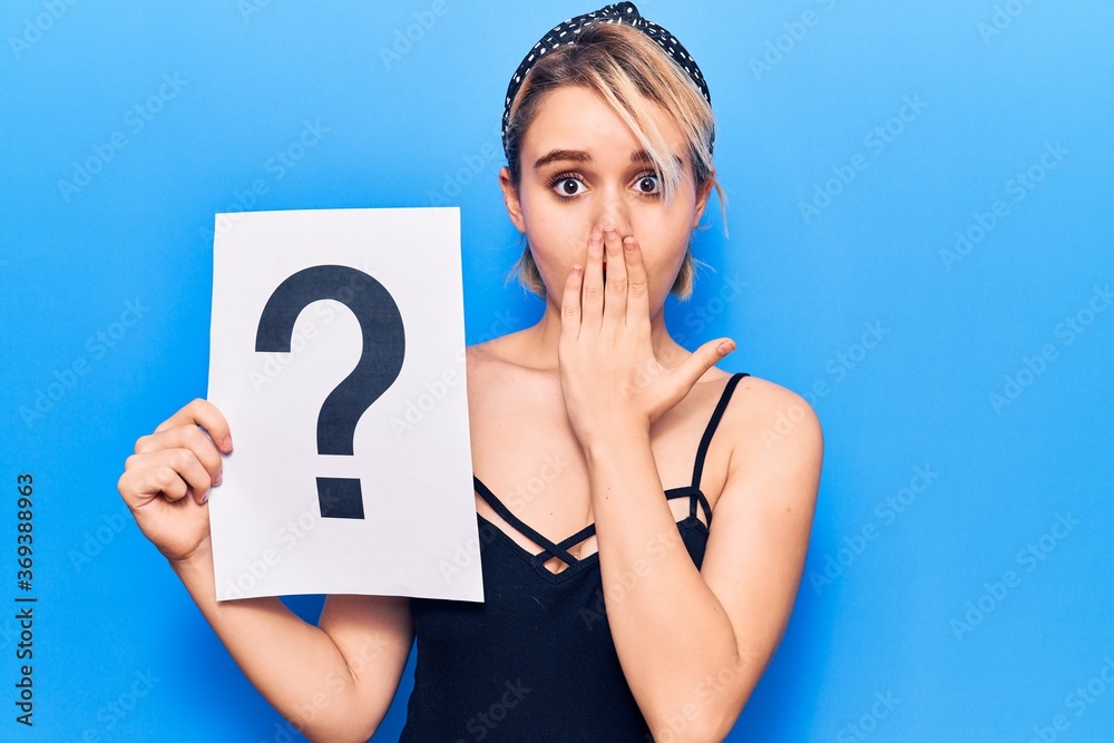 Young beautiful blonde woman holding question mark covering mouth with hand, shocked and afraid for mistake. surprised expression Young beautiful blonde woman holding question mark covering mouth with hand, shocked and afraid for mistake. surprised expression