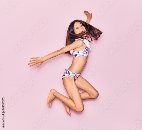 Adorable hispanic child girl on vacation wearing bikini surprised with open mouth. Jumping over isolated pink background