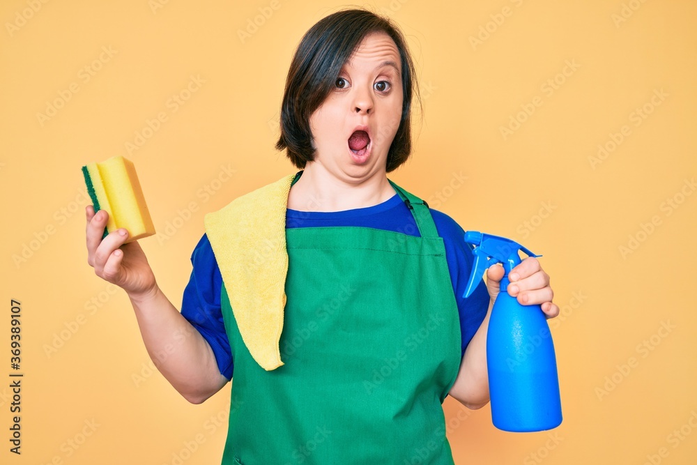 Brunette woman with down syndrome wearing apron holding scourer afraid and shocked with surprise and amazed expression, fear and excited face. Brunette woman with down syndrome wearing apron holding scourer afraid and shocked with surprise and amazed expression, fear and excited face.