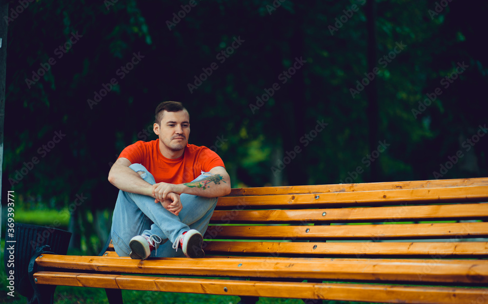Fototapeta premium Adult man resting on bench. Adult male in red t shirt and jeans sitting on bench and looking away while resting in park on summer day