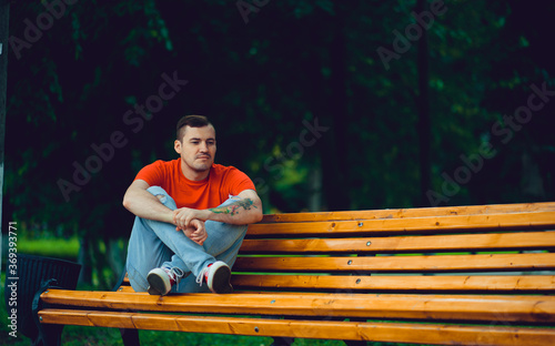 Adult man resting on bench. Adult male in red t shirt and jeans sitting on bench and looking away while resting in park on summer day