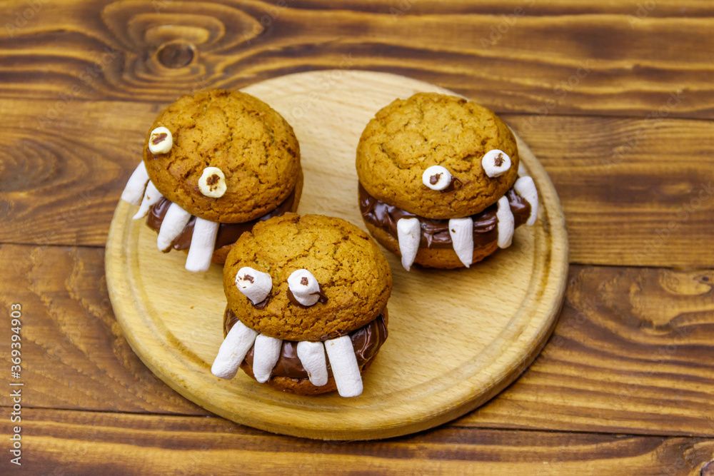 Fototapeta premium Halloween dessert: Toothed monsters made of oatmeal cookies, chocolate spread and marshmallows on a wooden table