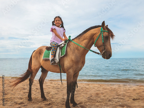 Portrait Asian child girl riding horse on a beach of Thailand in evening, background of sea and sky. Cute child girl with smiling face on brown horse, full body image.