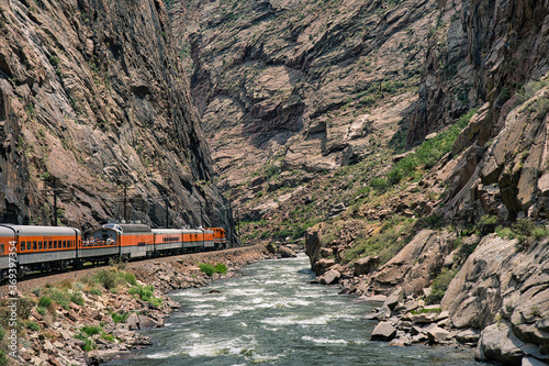 Train riding deep in the royal gorge beside the arkansas river in Colorado. Orange engine beside rushing blue water with rocky cliffs all around