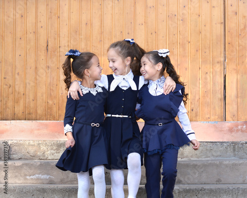 Three school friends have fun chatting down the steps. Elementary school students go amicably smiling at each other. Schoolgirls look at each other and smile. Girls in blue school clothes