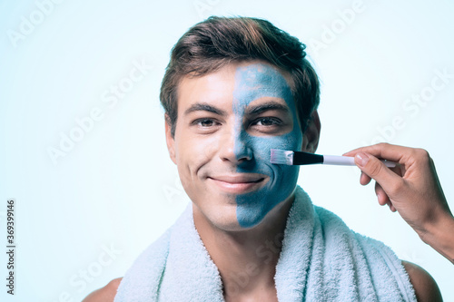 handsome man applying clay mask on face with brush while standing in bathrobe