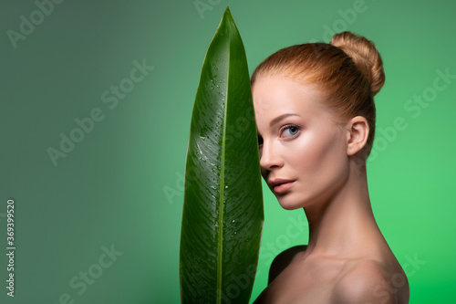 Portrait of healthy woman with half face is hidden behind a big tropical palm leaf on green background. Spa and wellness. Copy Space