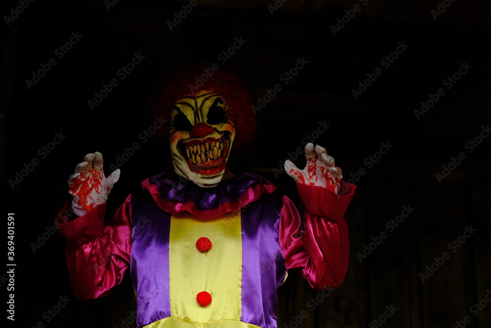 Halloween holiday. Creepy clown . Spooky clown in a bright overalls in the dark in a dark room.Autumn holidays in October. Festival and carnival concept. Horror and fear.Autumn seasonal holiday