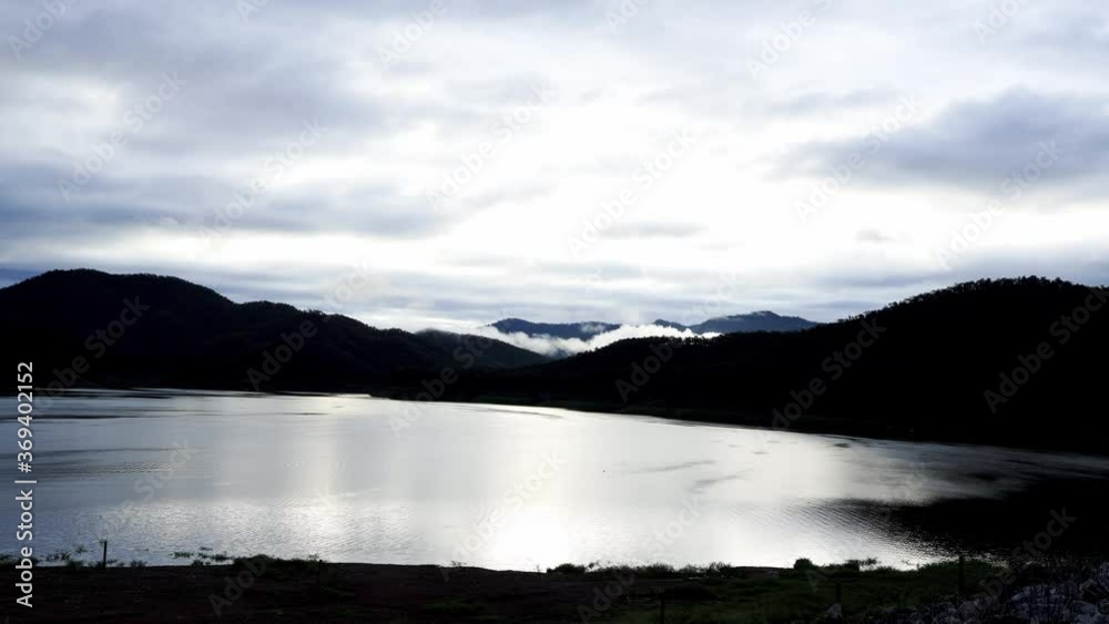 Time lapse of clouded sky after thunderstorm over the mountain range and the lake in the morning with rays of sunrise. A long-tailed boat running on the lake. .