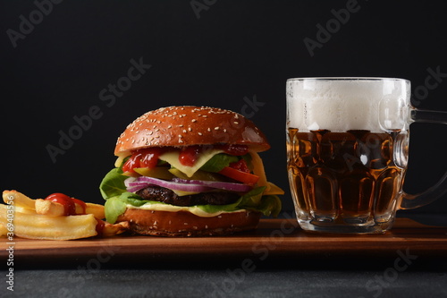 Homemade burger with grilled beef, cheese, onion, tomato, and green salad, sauce, with pickles and French fries,and a glass of beer in the background.