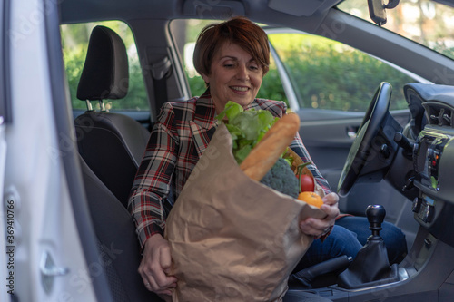 Beautiful middle age woman holding paper bag full of fresh vegetables and smiling. Going out from car after visiting super market. Carrying a healthy bag.
