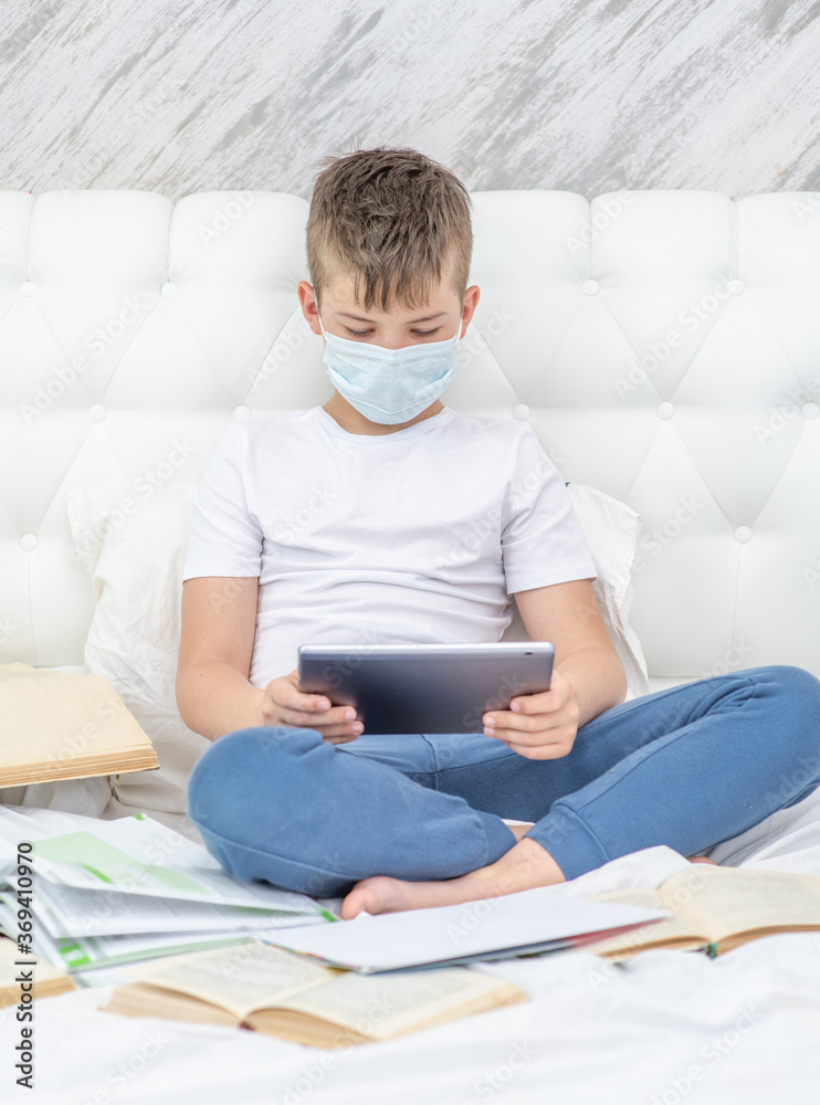 Smart boy wearing medical mask sits on the bed with tablet computer. Quarantine and coronavirus epidemic concept Smart boy wearing medical mask sits on the bed with tablet computer. Quarantine and coronavirus epidemic concept