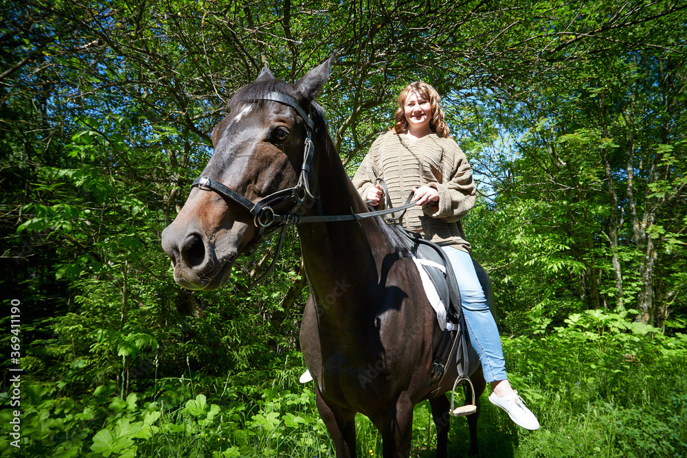 A fat girl and a brown horse in a Park on a Sunny day and green trees in the background. Young woman plus size rides a horse.