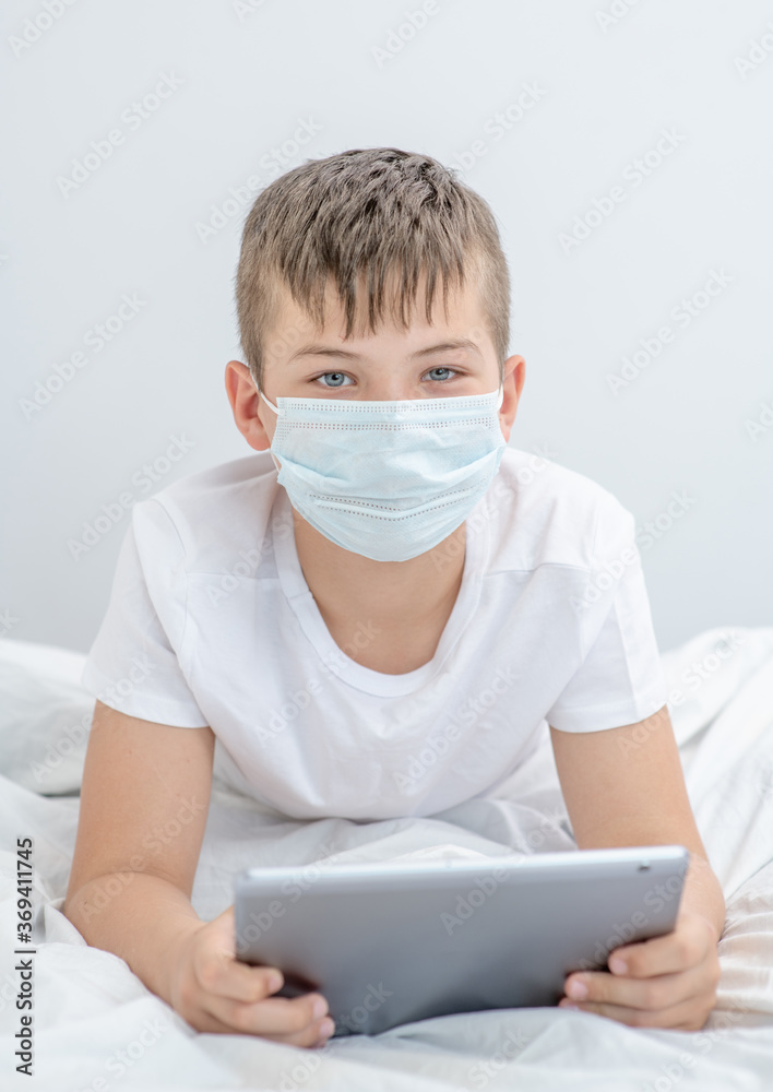 Portrait of a smart boy wearing medical mask lying on the bed with tablet computer. Quarantine and coronavirus epidemic concept