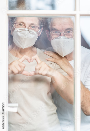 Elderly couple wearing protective face masks watch through their home window and show heart sign during the coronavirus epidemic