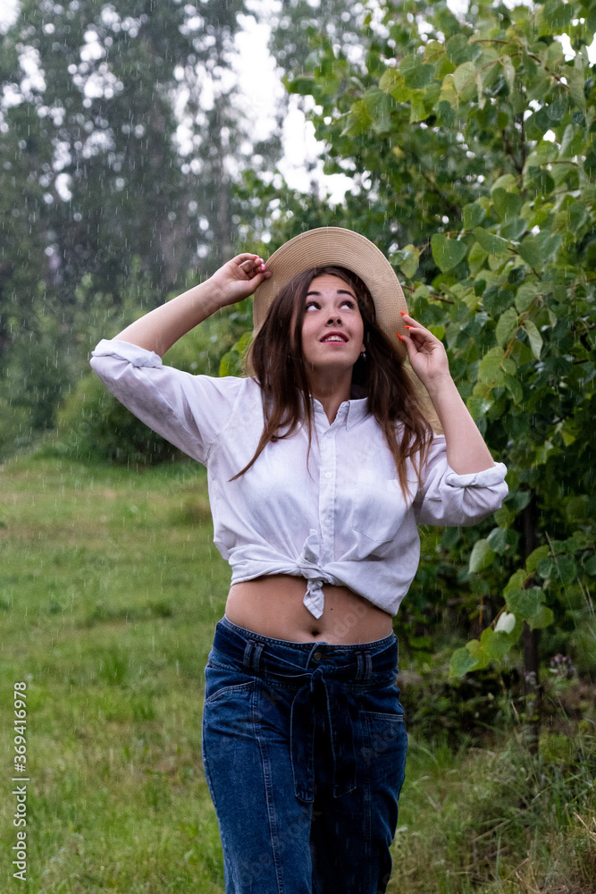 Summer warm rain. A young girl in a white shirt and blue jeans in the rain holds a hat on her head with her hands on a summer day against a background of greenery. Summer warm rain. A young girl in a white shirt and blue jeans in the rain holds a hat on her head with her hands on a summer day against a background of greenery.