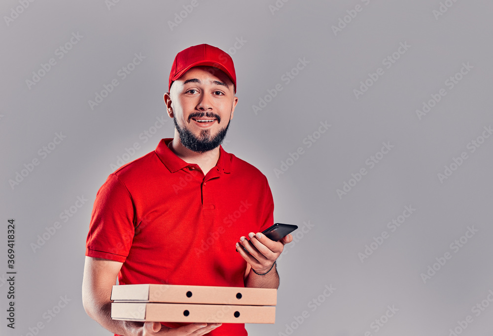 Bearded young delivery man in red T-shirt and cap with pizza boxes and with smartphone isolated on gray background. Fast home delivery.