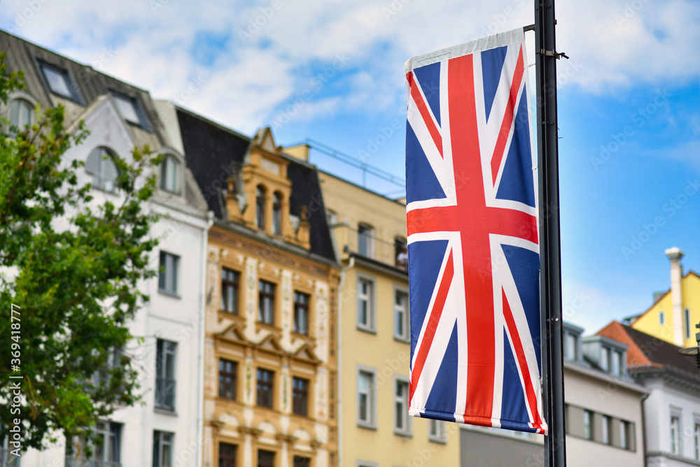 British Union Jack flag hanging on pole in front of blurry city street background with old buildings and blue sky British Union Jack flag hanging on pole in front of blurry city street background with old buildings and blue sky