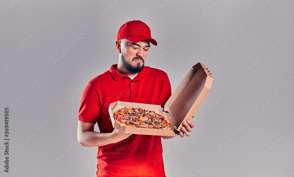 Young bearded delivery man in red uniform looks with disgust at pizza box isolated on gray background. Spoiled tasteless food. Young bearded delivery man in red uniform looks with disgust at pizza box isolated on gray background. Spoiled tasteless food.