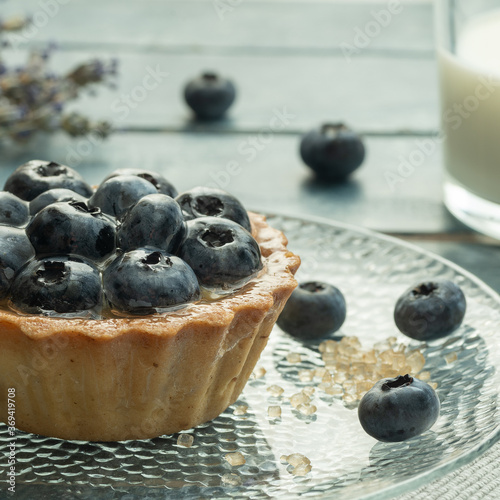 Blueberry tartlets served on a transparent plate with brown sugar. Glass of milk on a light blue wooden background.