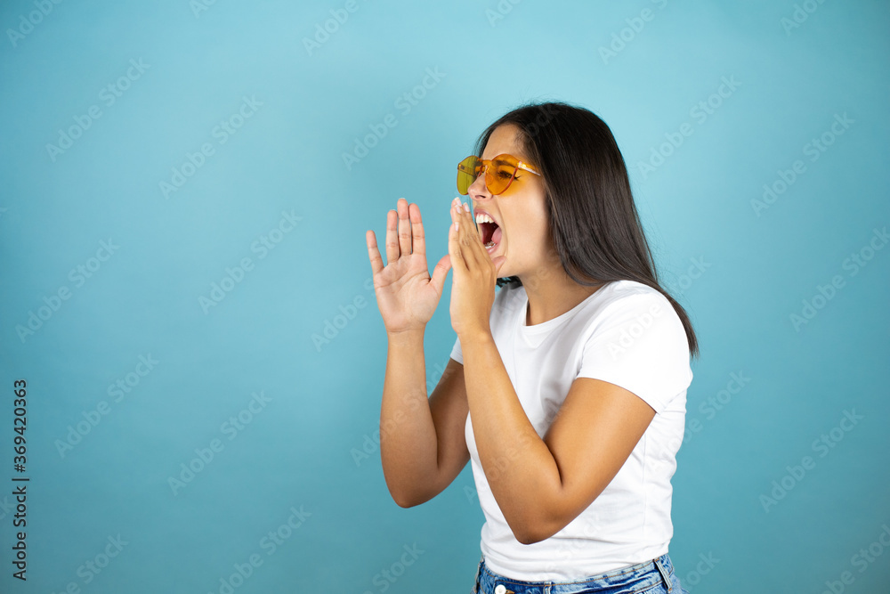 Young beautiful woman wearing sunglasses over isolated blue background shouting and screaming loud to side with hands on mouth Young beautiful woman wearing sunglasses over isolated blue background shouting and screaming loud to side with hands on mouth