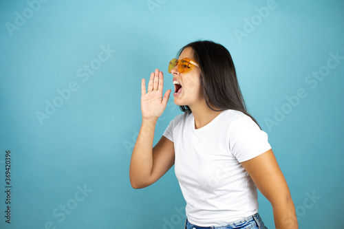 Young beautiful woman wearing sunglasses over isolated blue background shouting and screaming loud to side with hand on mouth