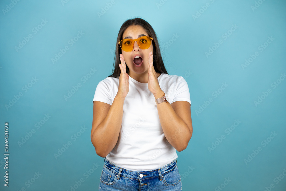 Young beautiful woman wearing sunglasses standing over blue isolated background with her hands over her mouth and surprised 